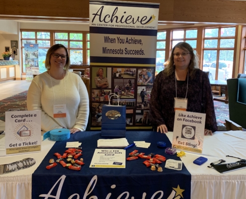 Nicky & Lynne stand behind the Achieve table at the Trainer & RBPD Summit.